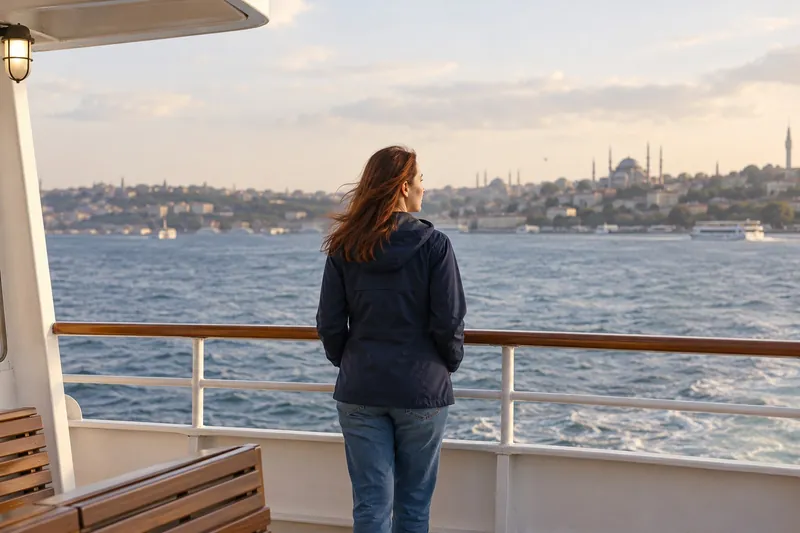 Kızıl-kumral saçlı bir kadın, vapurun açık güvertesinde yandan ve arkadan görünen bir açıyla Boğaz’a bakıyor.